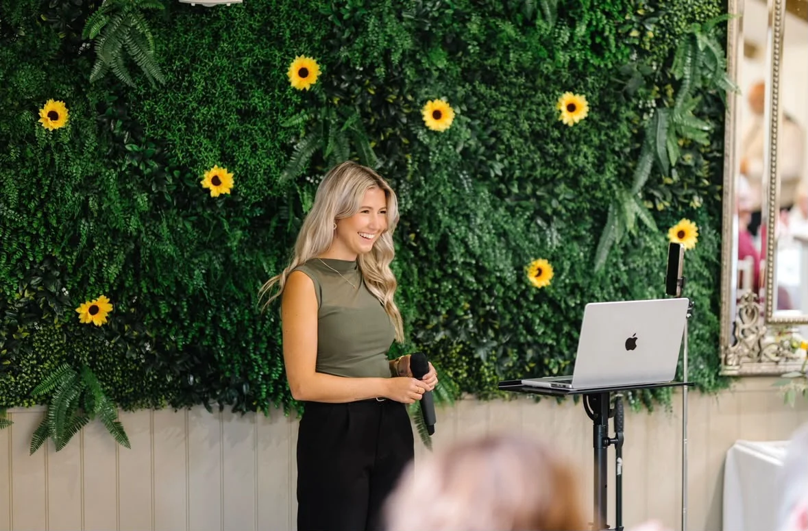 Caitie smiling beside her microphone with a floral wall behind her